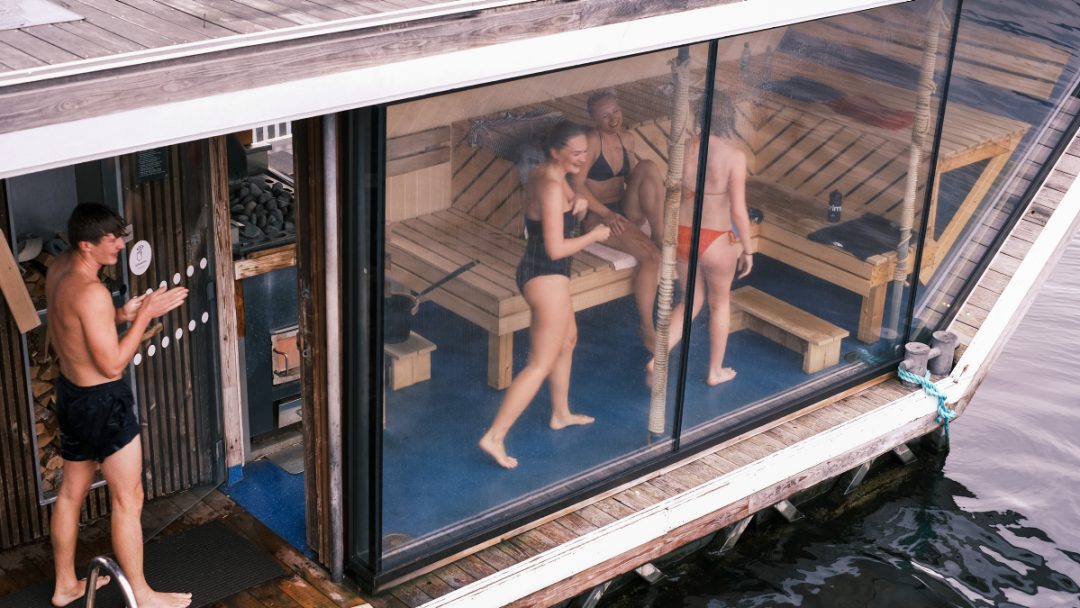 Folk i badedrakter nyter varmen fra en trebadstue ved vannet, mens en person utenfor tørker seg, kanskje lurer på noen nyttige badstutips.