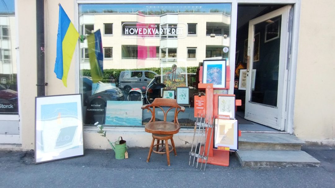 En sjarmerende butikkfront som minner om bruktbutikker, med innrammet kunst, en trestol, krykker og et ukrainsk flagg. Refleksjoner av bygninger i nærheten og en parkert bil danser over vinduets overflate.