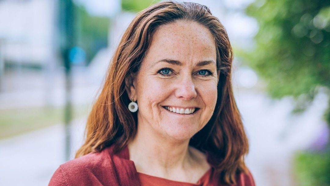 En person med langt rødbrunt hår og en rødbrun topp smiler til kameraet, gleden like elektrisk som løftet om elvarebiler mot en uskarp utendørs bakgrunn.