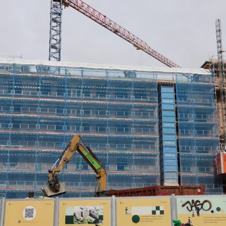 Byggeplassen har en delvis ferdigstilt bygning pakket inn i blått nett, med en kran og en gravemaskin som jobber aktivt. I forgrunnen viser en barriere informasjonsgrafikk om pågående anskaffelser og prosjektdetaljer.