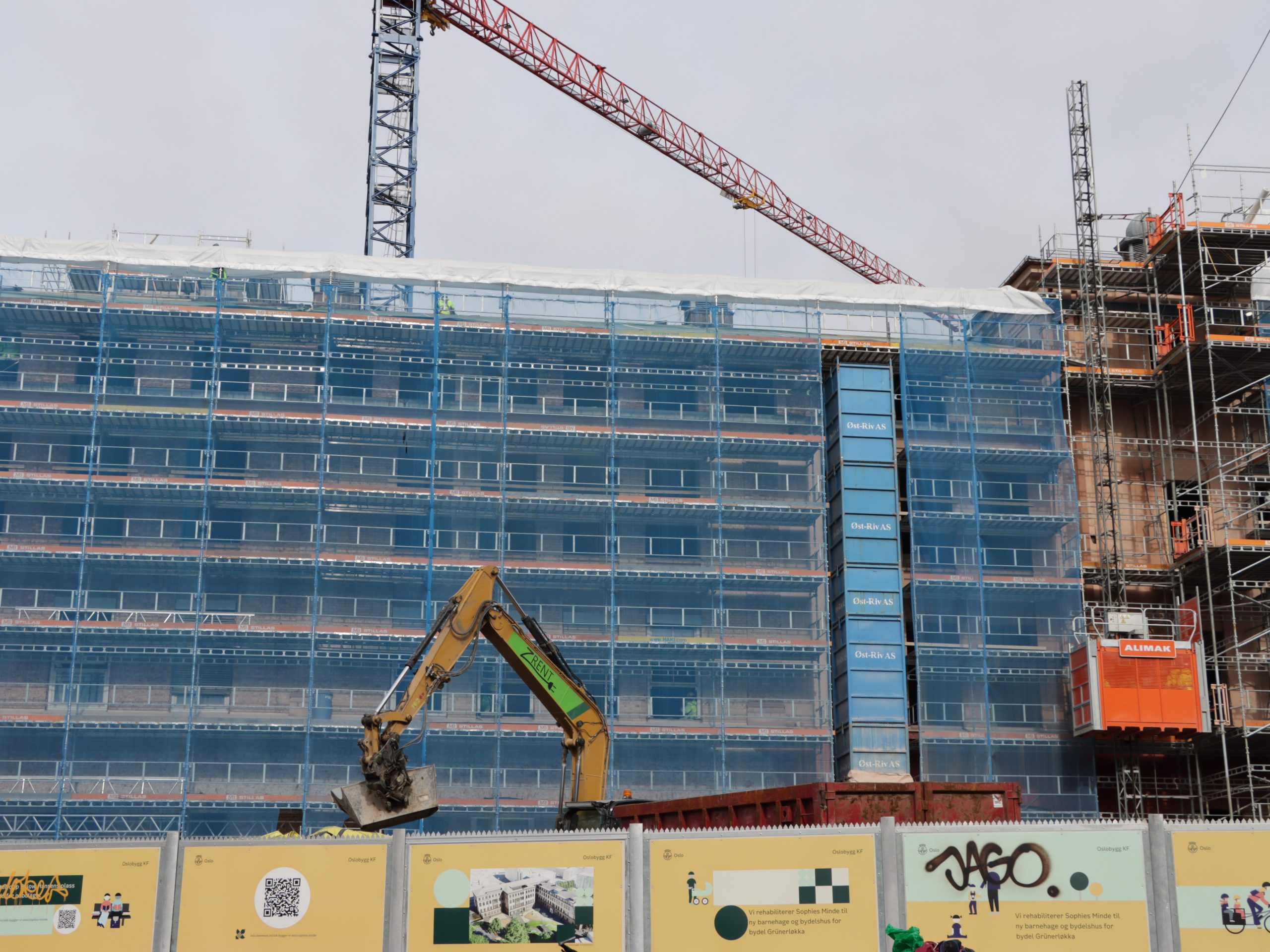 Byggeplassen har en delvis ferdigstilt bygning pakket inn i blått nett, med en kran og en gravemaskin som jobber aktivt. I forgrunnen viser en barriere informasjonsgrafikk om pågående anskaffelser og prosjektdetaljer.