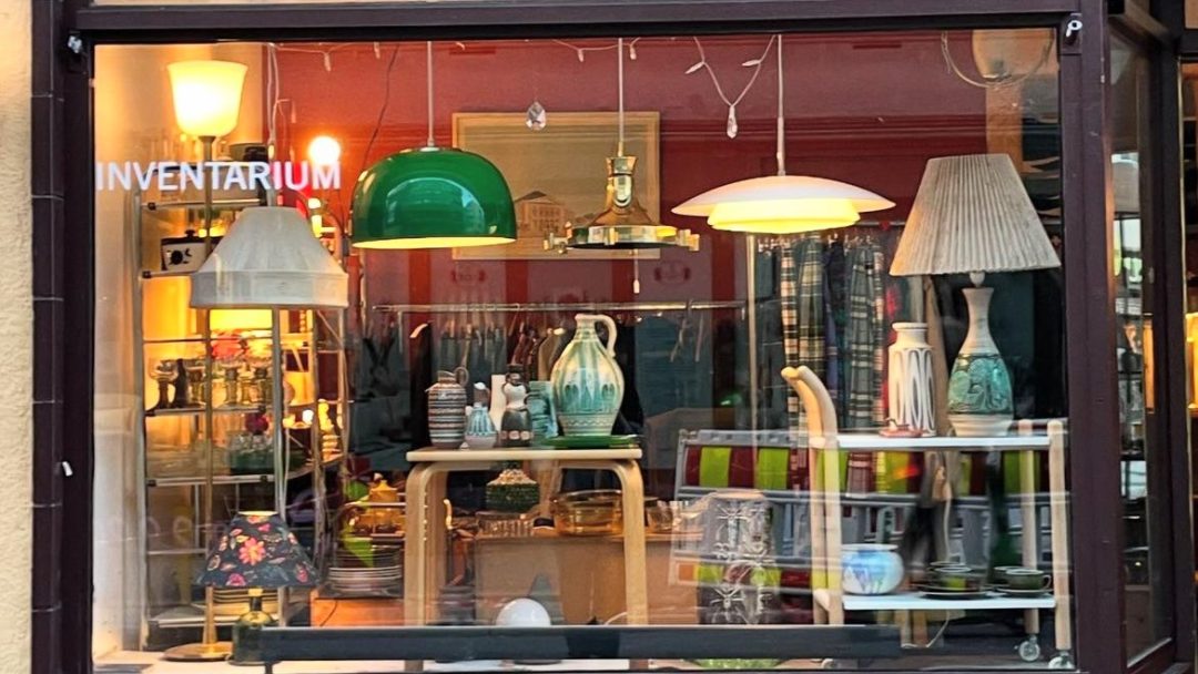 Utstillingsvinduet viser en sjarmerende samling av vintage bordlamper, vaser og stoler som ikke stemmer. Med skiltet Inventarium skiller denne perlen seg ut blant bruktbutikker for sine unike funn.