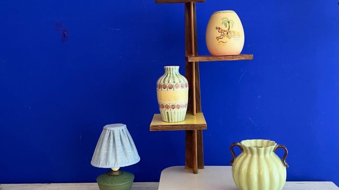 Assorterte vaser og en liten lampe, som minner om skatter funnet i bruktbutikker, står utstilt på en hvit overflate med en trehylle mot blå bakgrunn.