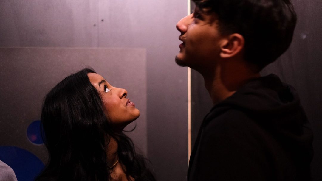 I det svakt opplyste rommet står to personer overfor hverandre. Gi, personen til venstre, ser opp på Julegave til høyre.