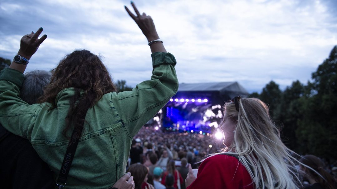 På en utendørskonsert svaier publikum i takt, med en person som gleder seg med fredstegn. Scenelys pulserer i bakgrunnen og skaper en levende, festlig atmosfære – en ekte gi til musikkelskere overalt.