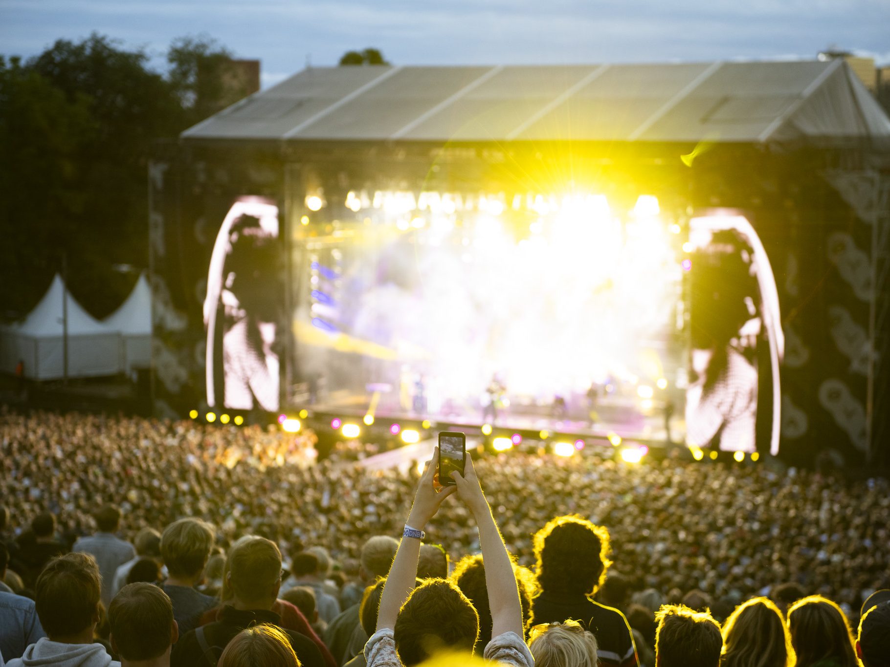 Et stort publikum deltar på en utendørskonsert med skarpe scenelys. En person i forgrunnen holder opp en telefon for å fange opptreden, kanskje tenker en video av Gi ville være en perfekt julegave.