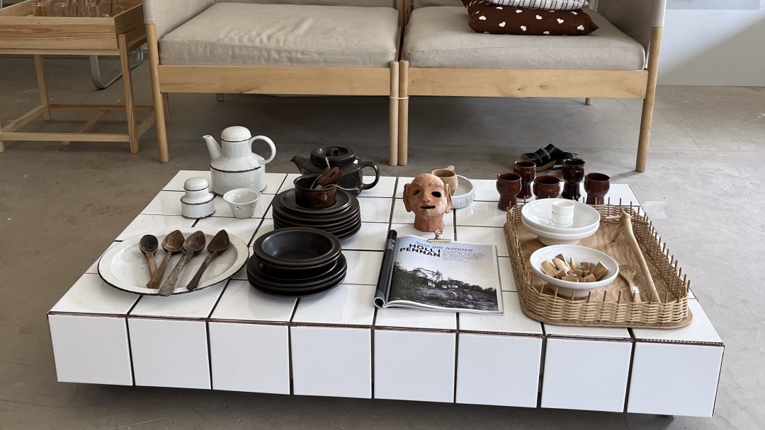 Et lavt, flislagt salongbord viser keramikkfat, treskjeer, en liten hundestatue og et magasin – skatter man kan finne i bruktbutikker. Bak den er en to-seters sofa med puter synlig.