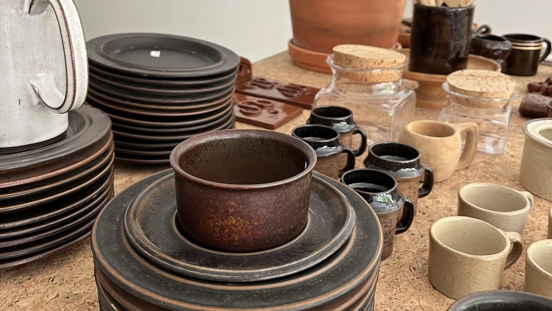 Diverse keramikkfat, krus og krukker er pent arrangert på en korkflate, som minner om funn fra sjarmerende bruktbutikker. Noen stykker er stablet, og treredskaper henger elegant i bakgrunnen.