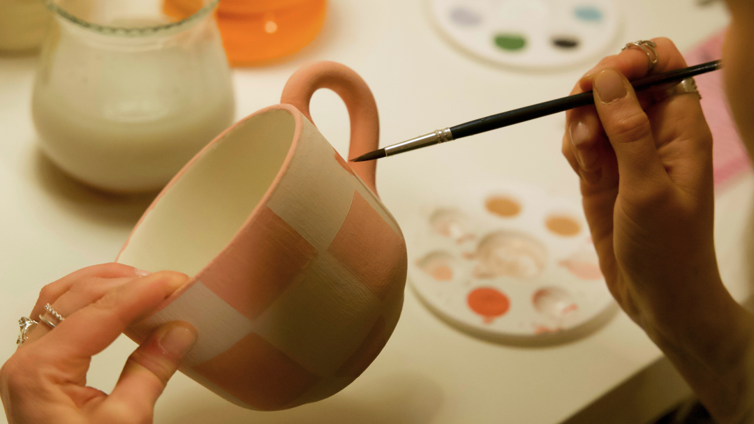 Person som maler en keramikkkopp med rosa firkantede mønstre med en liten pensel, kanskje lager den perfekte julegaven. En palett med forskjellige malingsfarger er synlig i bakgrunnen, og gir uendelige muligheter til å gi liv til kreativiteten.