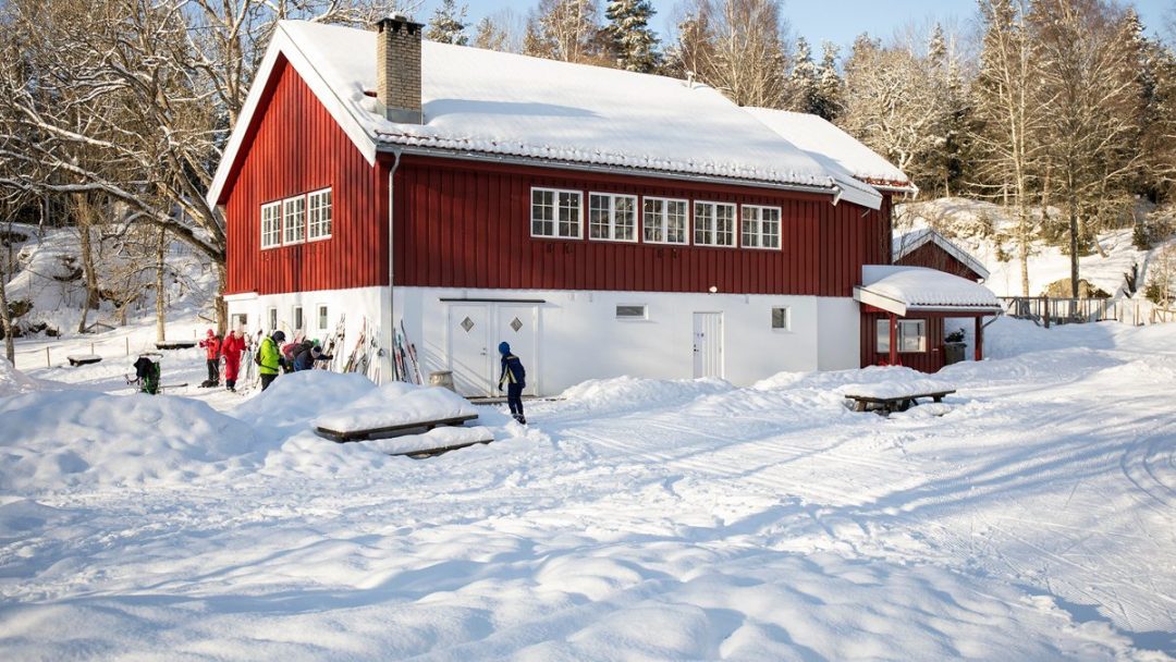 Et rødt trehus ligger i et snølandskap, sjarmen forsterkes av fnisende folk utenfor. Omgitt av trær og en snødekt bakke føles det som den perfekte scenen for utveksling av julegaver.
