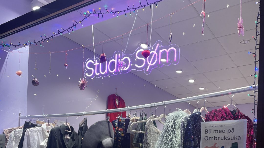 Butikkfronten, som minner om sjarmerende bruktbutikker, viser klær under et neon Studio Som-skilt. Ovenfor glimter dekorative lys og ornamenter innbydende.