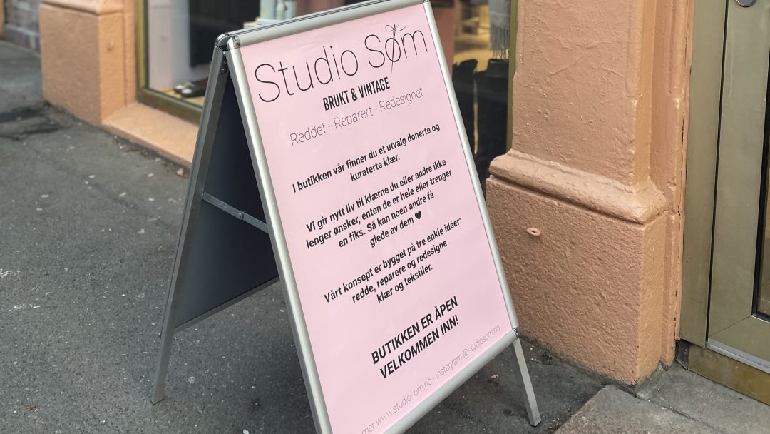 A-rammeskilt utenfor en butikk med en rosa plakat med informasjon om Studio Søm, en butikk som ligner på populære bruktbutikker, som tilbyr reparerte og resirkulerte gjenstander. På skiltet står det at butikken er åpen og ønsker besøkende hjertelig velkommen.