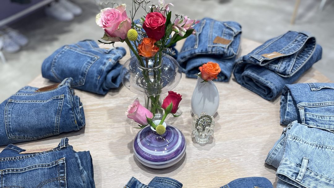 Brettede denimjeans fra lokale bruktbutikker er arrangert i en sirkel rundt et trebord, med vaser med fargerike blomster som lysner opp midten.