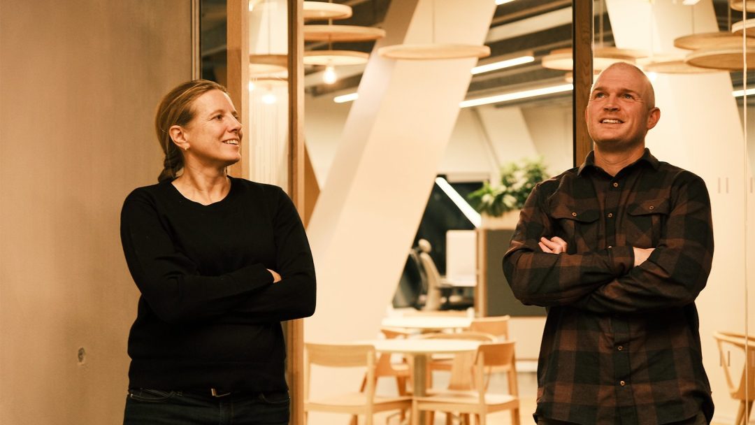 To personer med armer i kors står og ser oppover i Vertikal Nydalens kontorlokale, med tremøbler og taklamper.