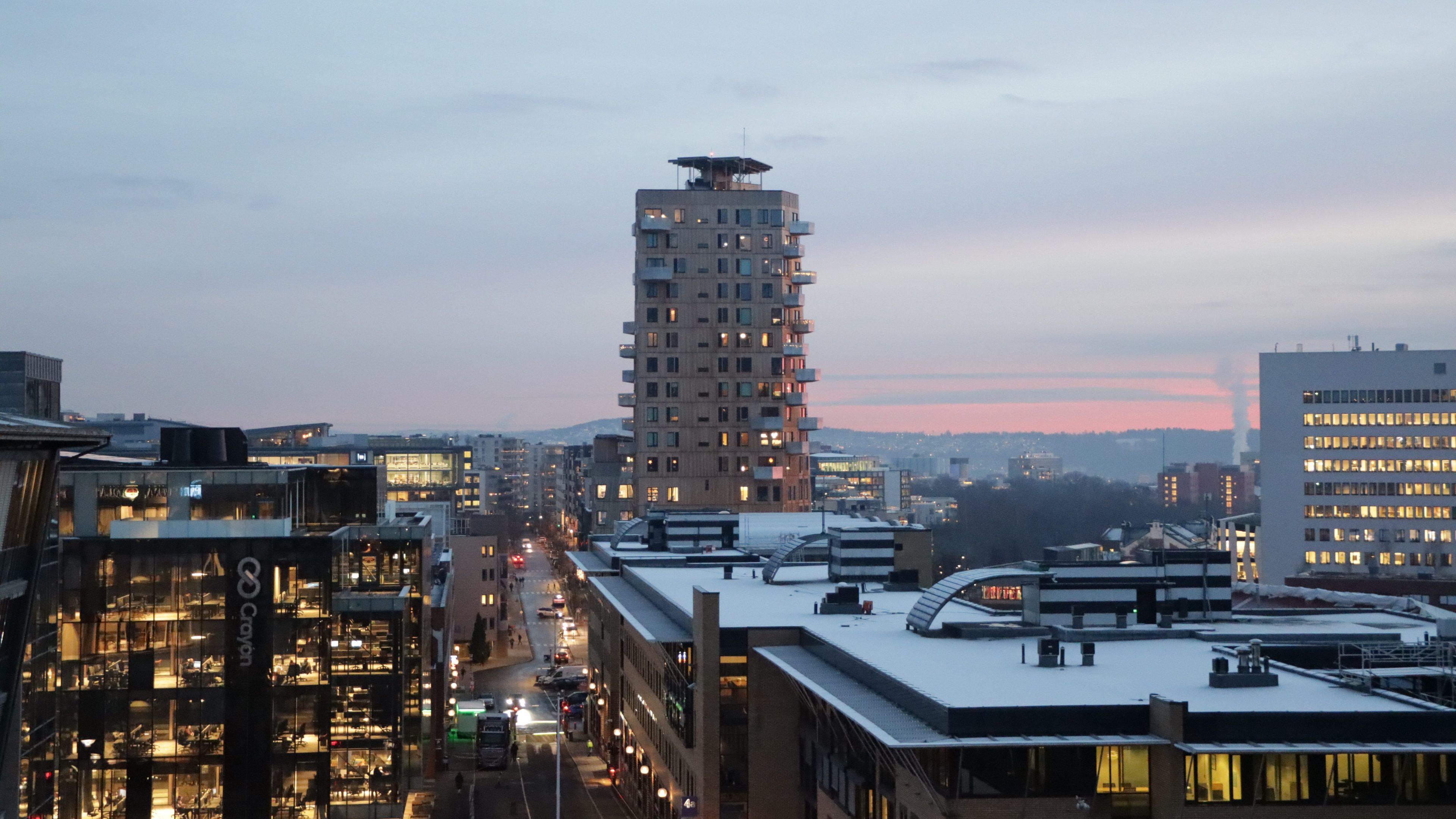 I skumringen avslører bybildet Vertikal Nydalen, et høyt, moderne bolighus plassert blant kontortårn. Gater med biler surrer mens lys skimrer i vinduene, og himmelen lyser av rosa fargetoner.