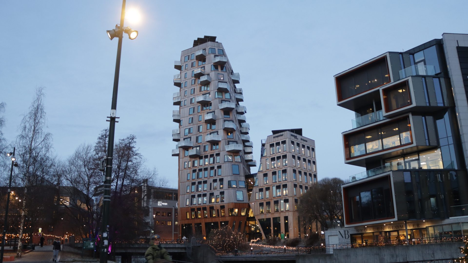 De moderne, unikt formede bygningene i Vertikal Nydalen med kantete design og mange vinduer står høyt mot en skumringshimmel. Gatelys lyser opp scenen, mens trær svaier forsiktig i forgrunnen.
