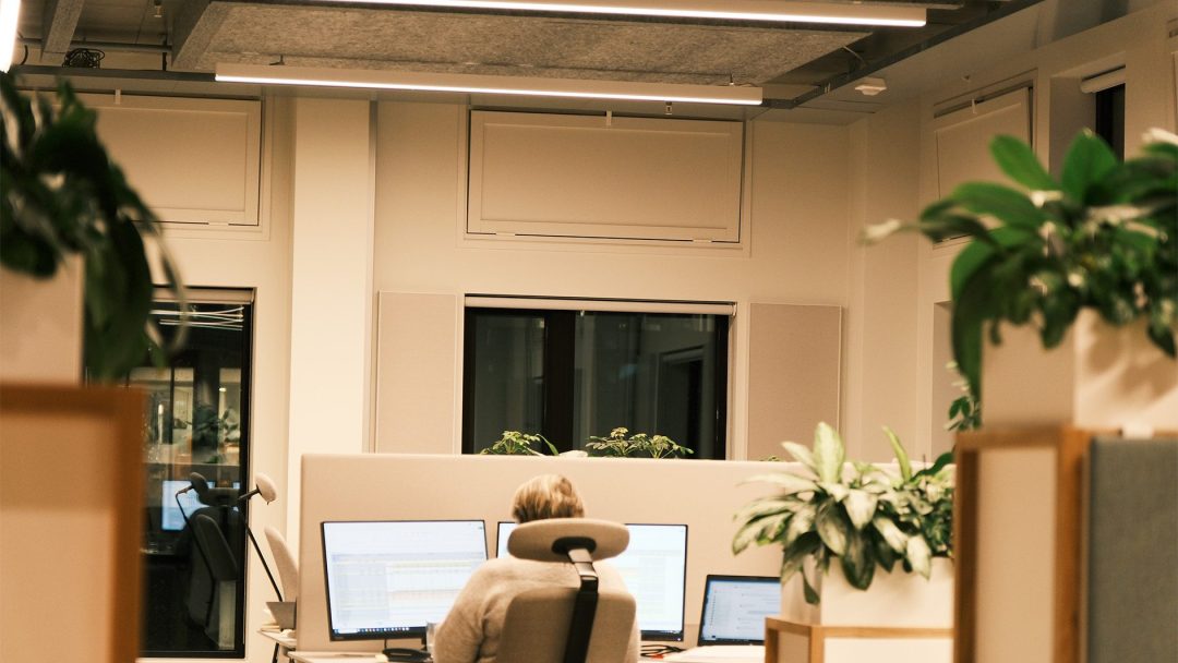 På det travle kontoret til Vertikal Nydalen sitter en person ved et skrivebord med tre monitorer, omgitt av potteplanter og beige skillevegger.