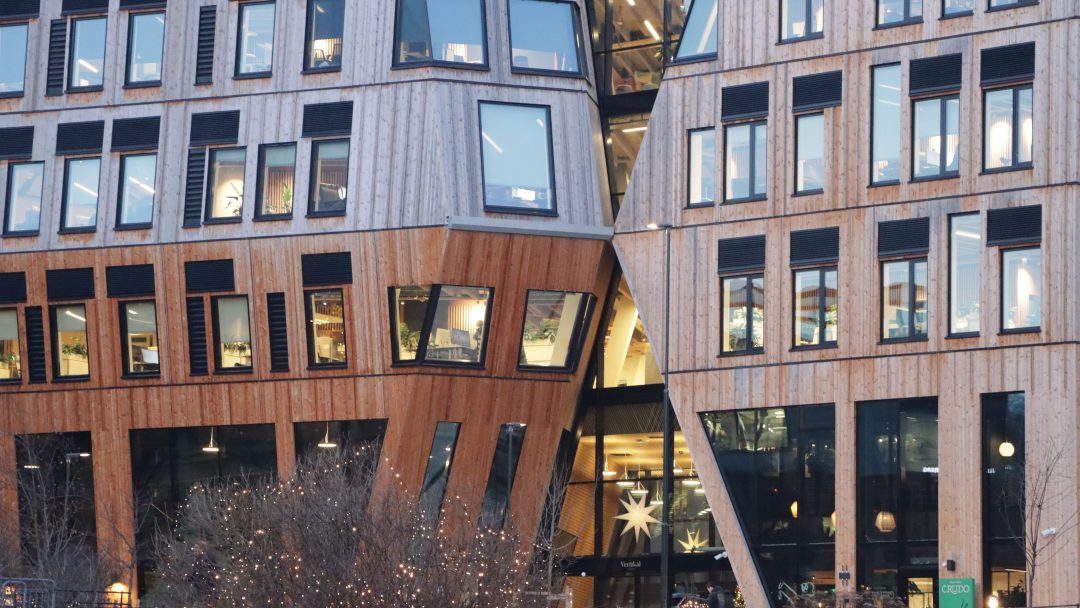 Vertikal Nydalen har to moderne, kantete bygninger med store vinduer, kledd i tre og glass, skjev mot hverandre. Trær og lyssnorer pryder elegant forgrunnen, og skaper en rolig atmosfære.