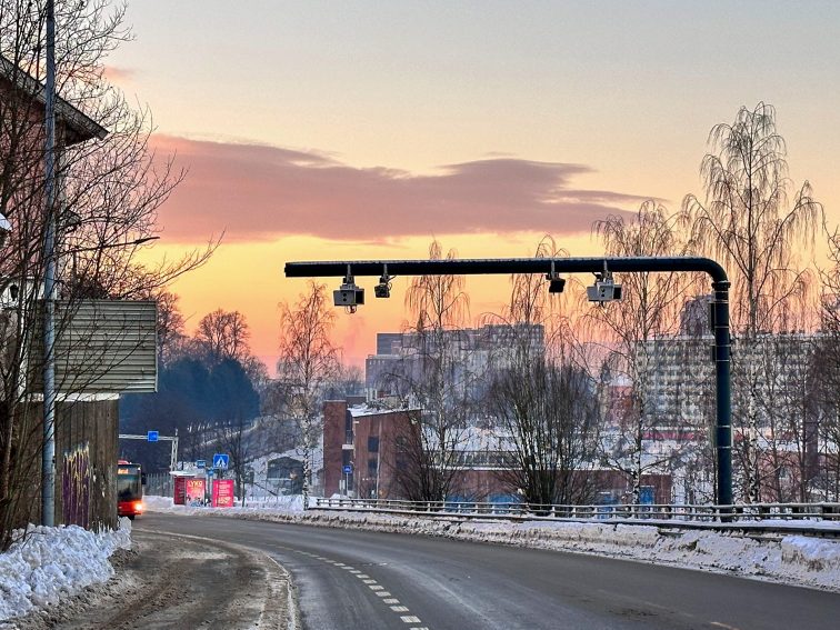 Snødekt byvei ved solnedgang med trafikkkameraer over hodet og bygninger i det fjerne. Elvarebiler glir stille langs gaten omkranset av nakne trær, og legger til et snev av modernitet til den rolige vinterscenen.