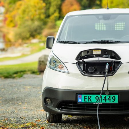 Et hvitt elektrisk kjøretøy lader utendørs, med en kabel plugget i fronten. Scenen fremhever fordeler ved å kjøre elvarebil i Oslo, satt mot et bakteppe av trær og en vannmasse.
