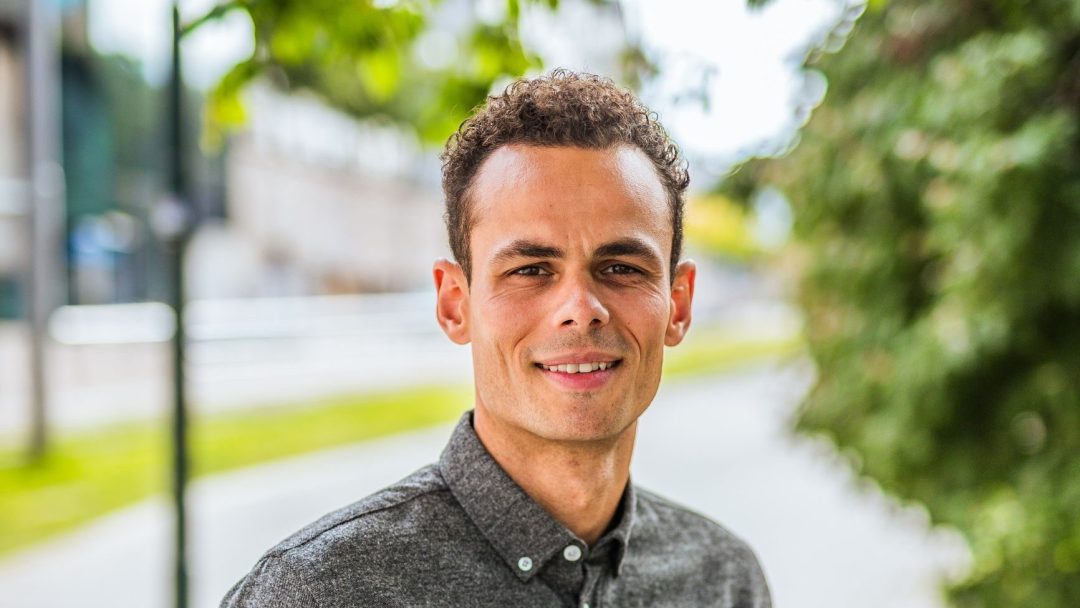 En person med kort, krøllete hår smiler utenfor, iført en grå skjorte med knapper som tilfører et snev av eleganse. I den uskarpe bakgrunnen skaper grøntområder og en sti en rolig scene, omtrent som atmosfæren til taxiladere.