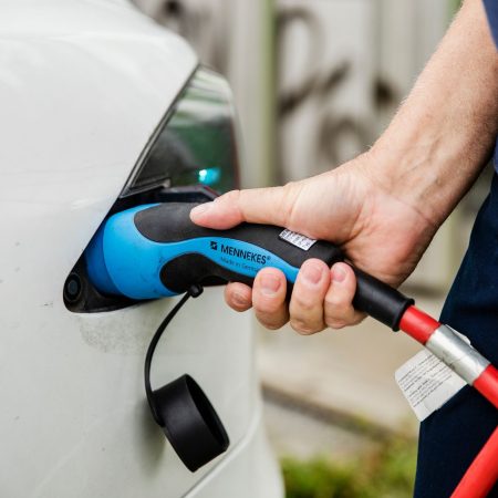En hånd kobler en blå elbil-lader inn i ladeporten på et hvitt elektrisk kjøretøy, med den røde kabelen til taxiladeren synlig.
