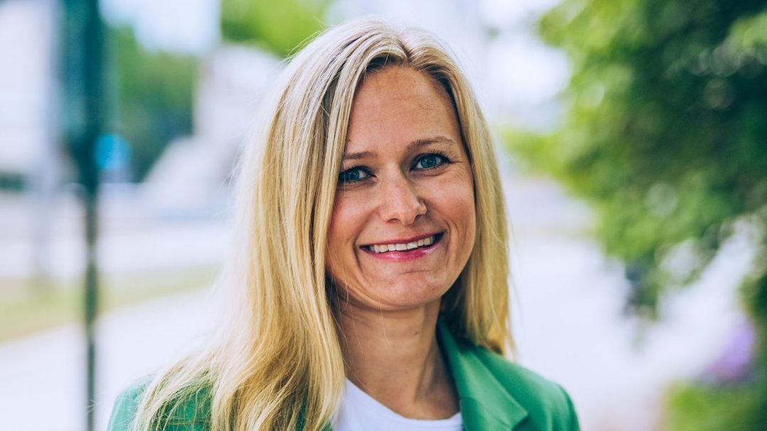 En kvinne med langt blondt hår og en grønn blazer smiler utendørs, omgitt av trær og en sløret gate fylt med elvarebiler i bakgrunnen.