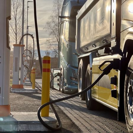 Under en klar blå himmel står tungtransport elektriske lastebiler i kø ved en ladestasjon, hver perfekt koblet sammen med solide kabler, klare til å starte veien videre.