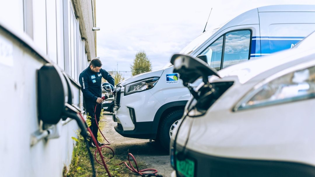 En person håndterer anskaffelsen av en elektrisk kabel, kobler den til en hvit varebil på parkeringsplassen, mens en annen varebil står parkert i nærheten.
