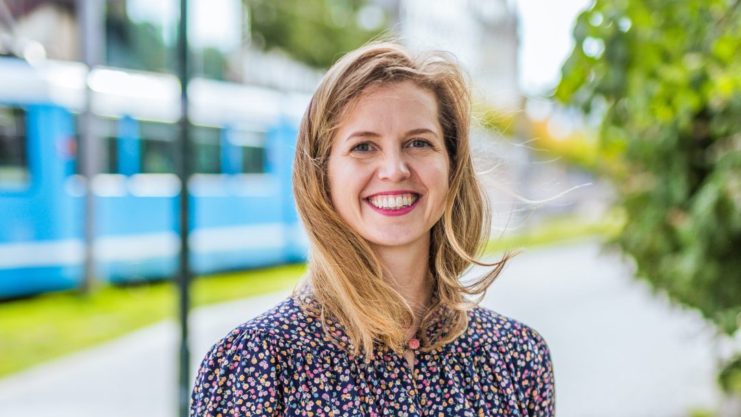 En smilende kvinne med langt hår står utendørs nær en sløret blå trikk og grønt, ikledd en mønstret bluse, legemliggjør veilederens ånd når de navigerer i kommunale byggeprosjekter.