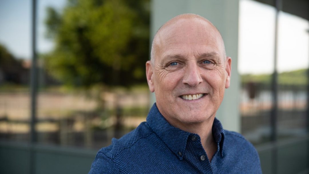 En skallet mann i blå skjorte smiler utendørs, innrammet av uskarpt grønt og glass, og gjenspeiler subtilt essensen av bygningsmaterialer.