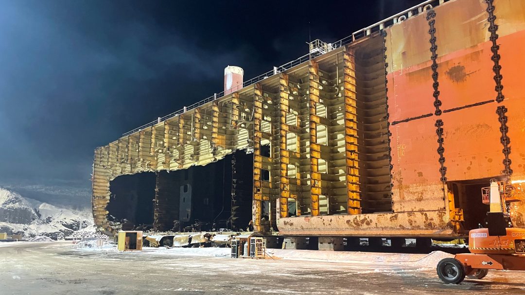 Det store, rustne skipsskroget, med sin indre struktur synlig, hviler på et snødekt industriområde om natten. Bygningsmaterialer lå spredt rundt mens den iskalde vinden hvisker gjennom stålbeina til denne maritime kjempen.