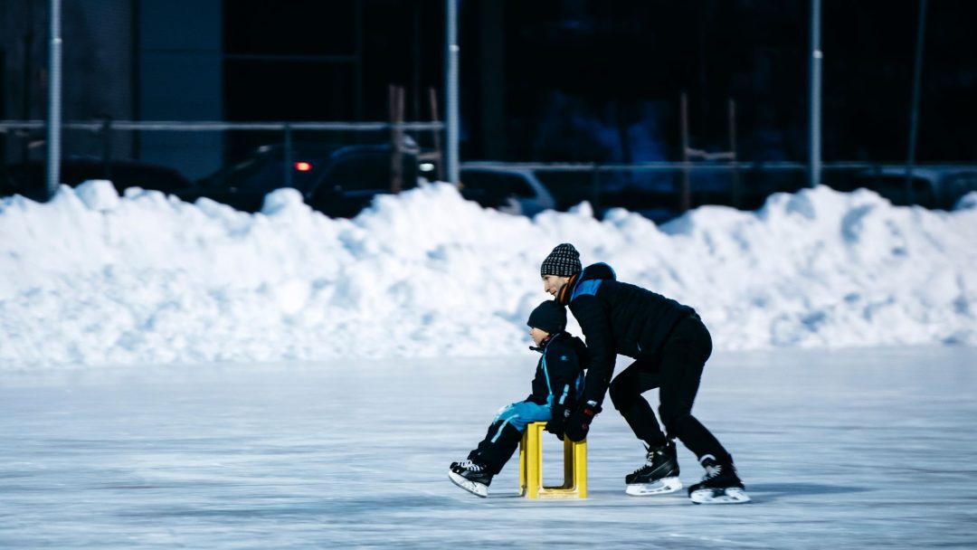 En voksen skater glir grasiøst over den utendørs skøytebanen og dytter et barn som sitter på en gul boks. Scenen fanger den gledelige essensen av skøyter med snø pent stablet i bakgrunnen, og skaper et pittoresk vinterlandskap.
