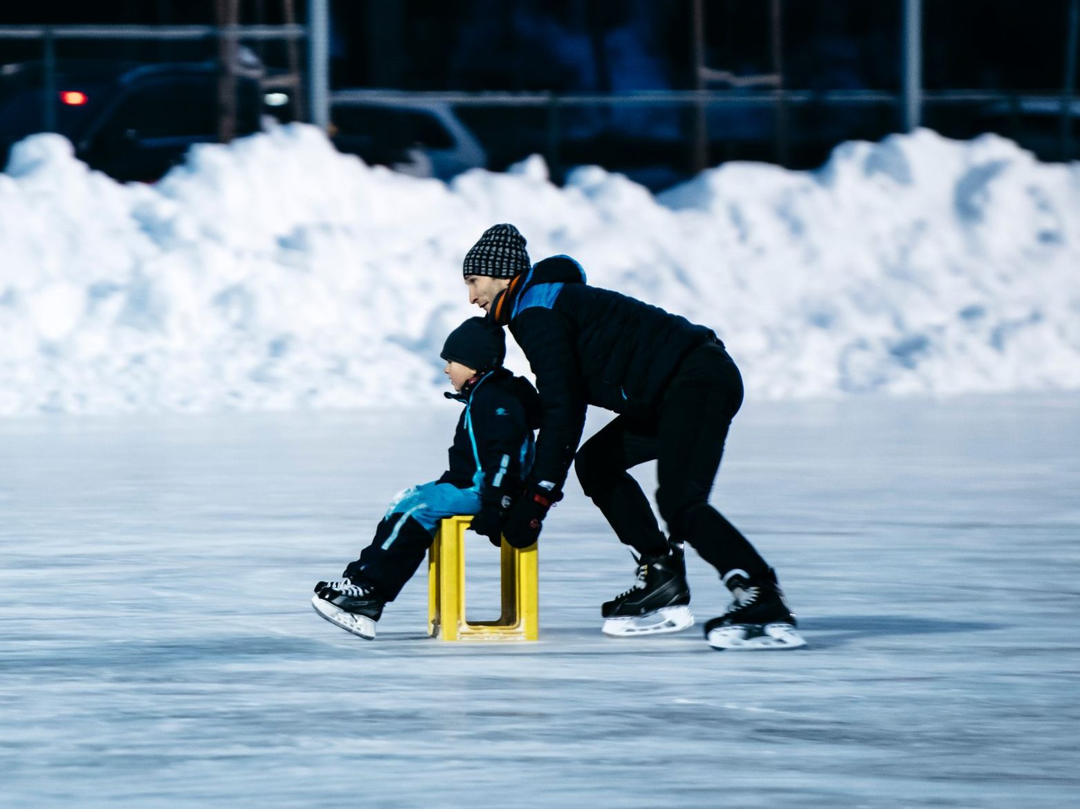 En voksen guider et barn mens de skøyter på isen, ved hjelp av en gul stol som støtte på en utendørs bane, med snø som forsiktig dekker bakgrunnen.