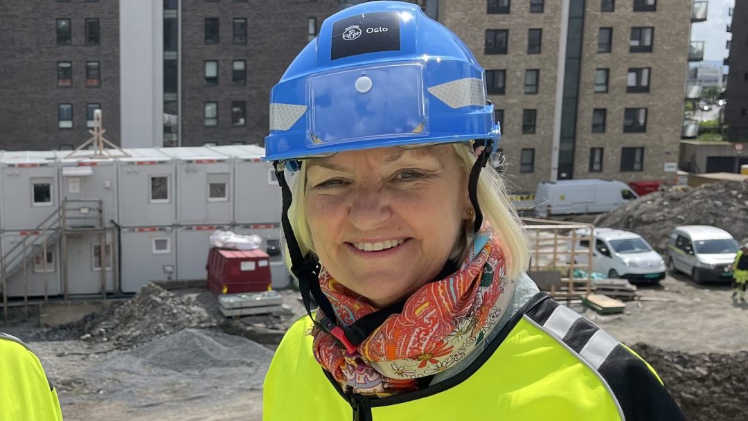 En kvinne i blå hjelme og gul sikkerhetsjakke står på en byggeplass, omgitt av stabler med bygningsmaterialer, med bygninger som reiser seg majestetisk i bakgrunnen.