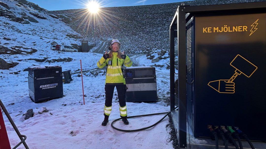 En person i en synlig jakke og hjelm står i et snødekt terreng, i nærheten av elektrisk utstyr, med solen skinnende i bakgrunnen.