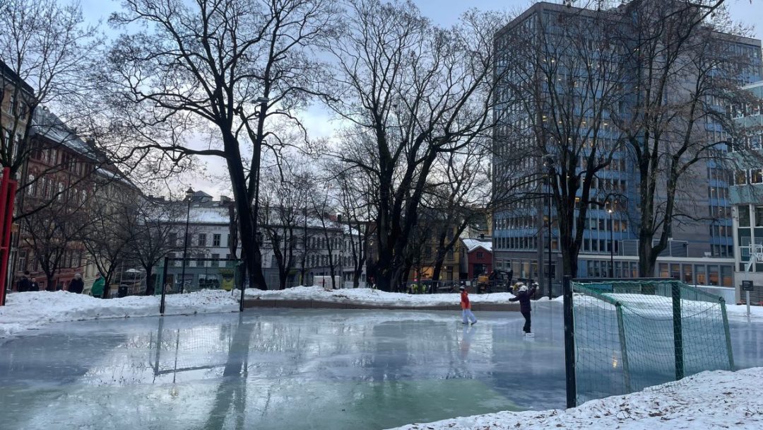To personer glir grasiøst på skøyter over en liten skøytebane, omgitt av snøstøvede trær og urbane bygninger i bakgrunnen.