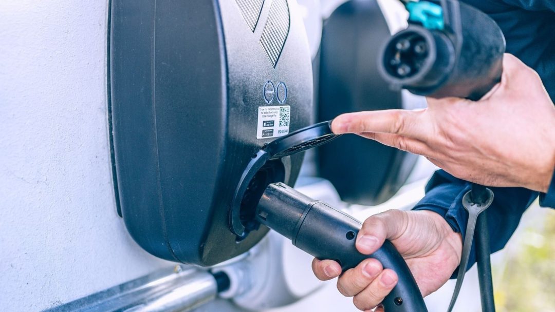 En person kobler en kabel til en ladestasjon for elektriske kjøretøy montert på en vegg, og viser en konkret klimatiltak innsats for å støtte bærekraftig transport.