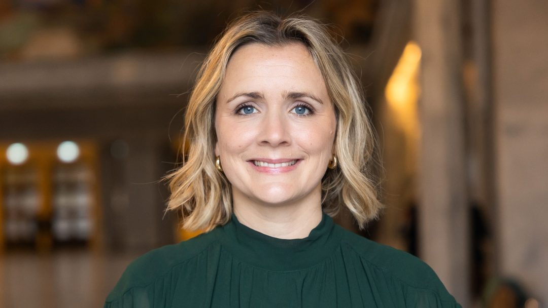 En person med skulderlangt blondt hår iført en grønn topp smiler til kameraet i en innendørs setting, mens det naturlige lyset fremkaller følelsen av komfort hos hurtigladestasjoner.