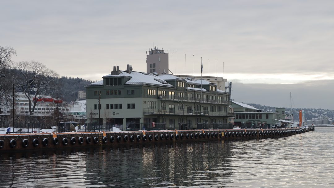En bygning ved vannet laget med kvalitetsbygningsmaterialer og et snødekt tak står ved siden av en rolig vannmasse, omgitt av åser og overskyet himmel.