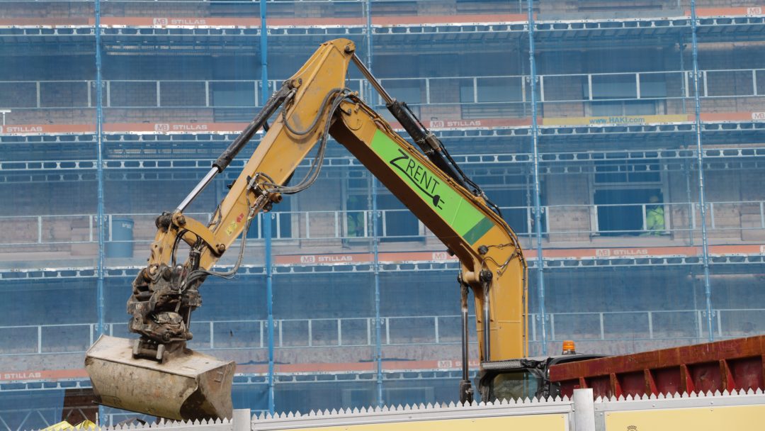 En gravemaskinarm løfter et lass ved et kommunalt byggeprosjekter område med delvis overbygd bygning i bakgrunnen.