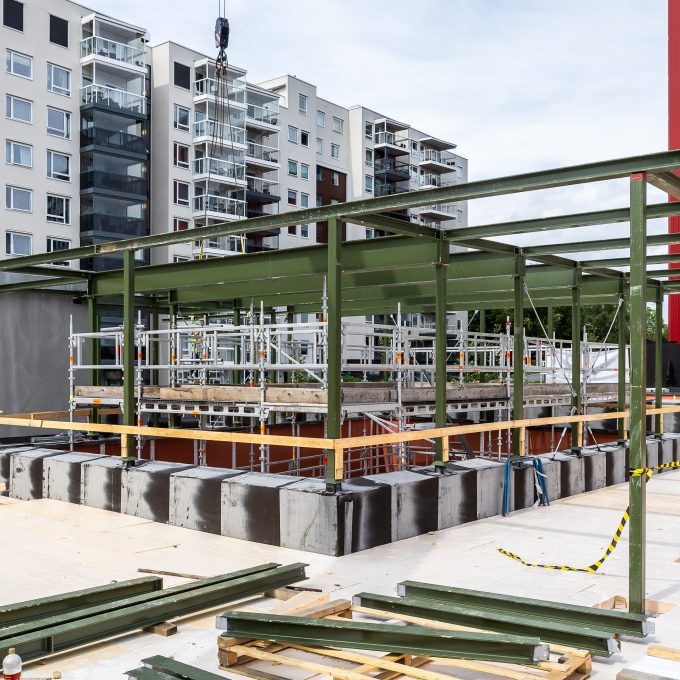 En byggeplass med delvis bygget struktur viser solide bygningsmaterialer, med stålbjelker og betongfundamenter. Moderne leilighetsbygg skaper en imponerende skyline i bakgrunnen.