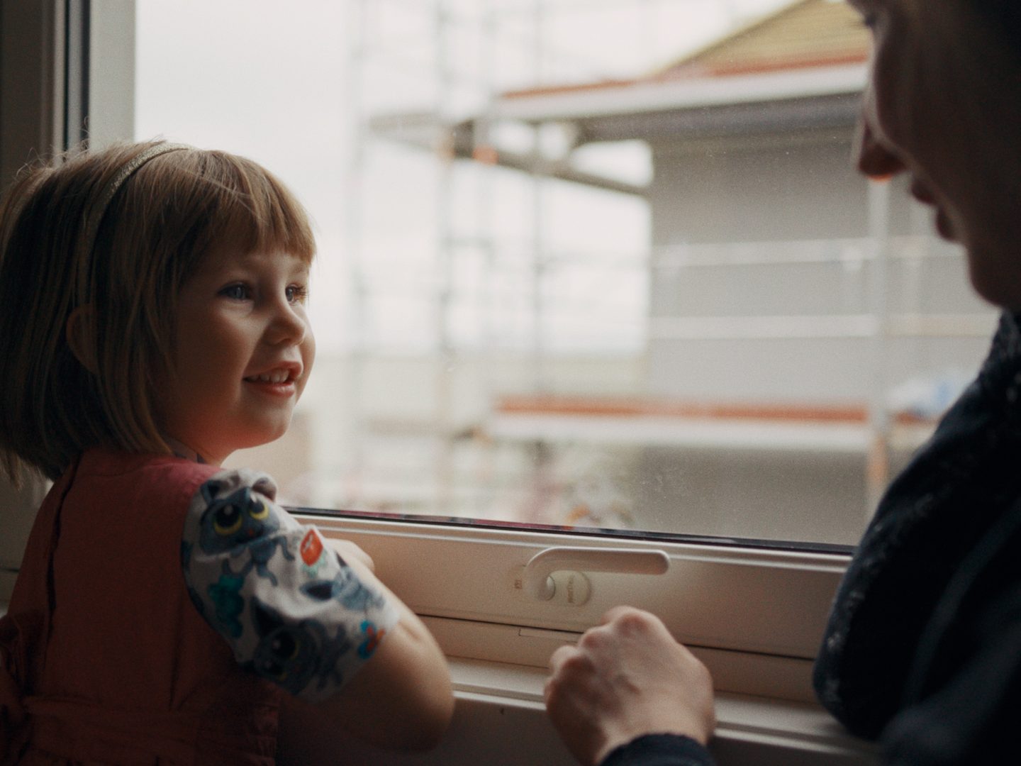 Et lite barn og en voksen ser ut av vinduet. Barnet, iført en fargerik skjorte, smiler mens de begge ser på en bygning under bygging. Scenen er innrammet av energieffektive vinduer, som antyder moderne bærekraft.