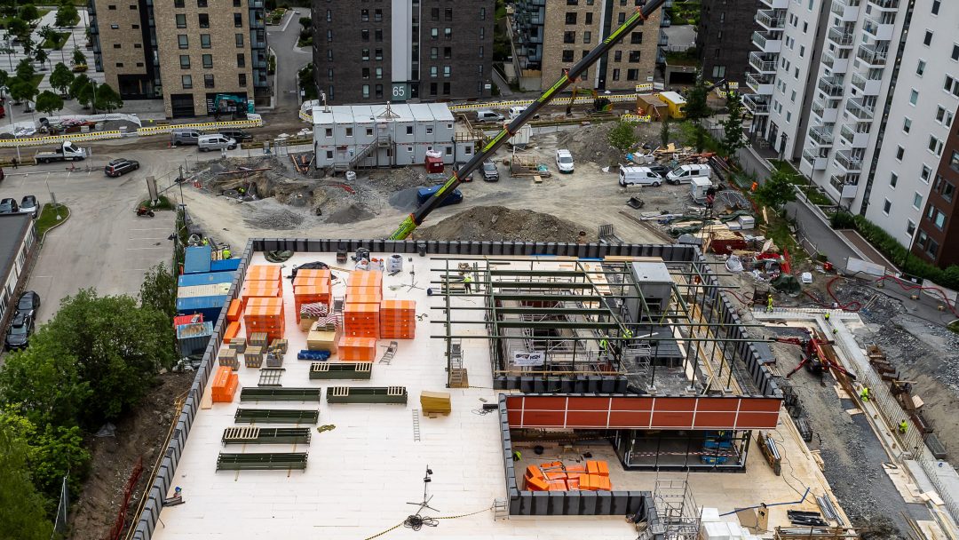 Luftfoto av en byggeplass yrende av maskiner, bygningsmaterialer og flittige arbeidere. Omkringliggende bygninger og veier smelter sømløst inn i scenen.