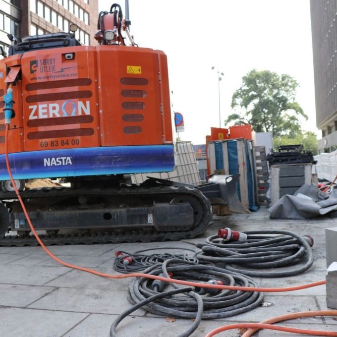 En travel byggeplass har en oransje gravemaskin og opprullede kabler på bakken, alt omgitt av høye bygninger. Den sømløse anskaffelsen av materialer sikrer at alle elementer er på plass for konstruksjonens suksess.