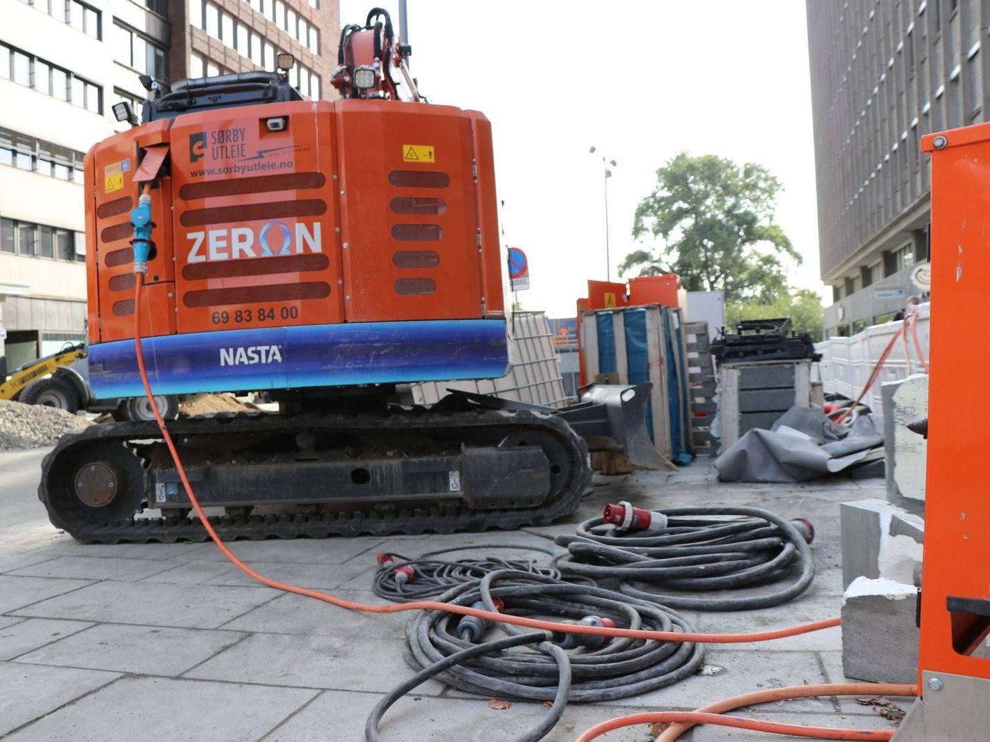 En travel byggeplass har en oransje gravemaskin og opprullede kabler på bakken, alt omgitt av høye bygninger. Den sømløse anskaffelsen av materialer sikrer at alle elementer er på plass for konstruksjonens suksess.