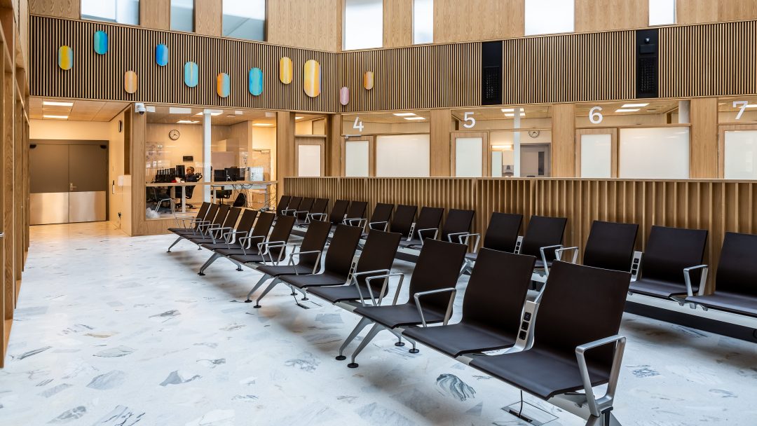 Det tomme venteområdet har rader med svarte stoler, en skillevegg i tre og nummererte servicevinduer, alt satt opp mot fargerike veggdekorasjoner i et moderne rom som viser innovative bygningsmaterialer.