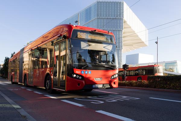 En rød bybuss glir langs gaten ved siden av en moderne glassbygning under klar himmel, samtidig som den bidrar til klimagassutslippene.