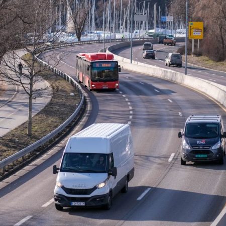 Oversikt over en vei med en hvit varebil, en grå varebil og en rød buss som bidrar til klimagassutslippene. To syklister sykler på en sti ved siden av veien da seilbåter er synlige i bakgrunnen.