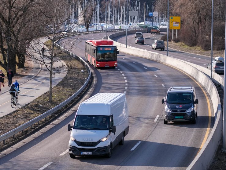 Oversikt over en vei med en hvit varebil, en grå varebil og en rød buss som bidrar til klimagassutslippene. To syklister sykler på en sti ved siden av veien da seilbåter er synlige i bakgrunnen.
